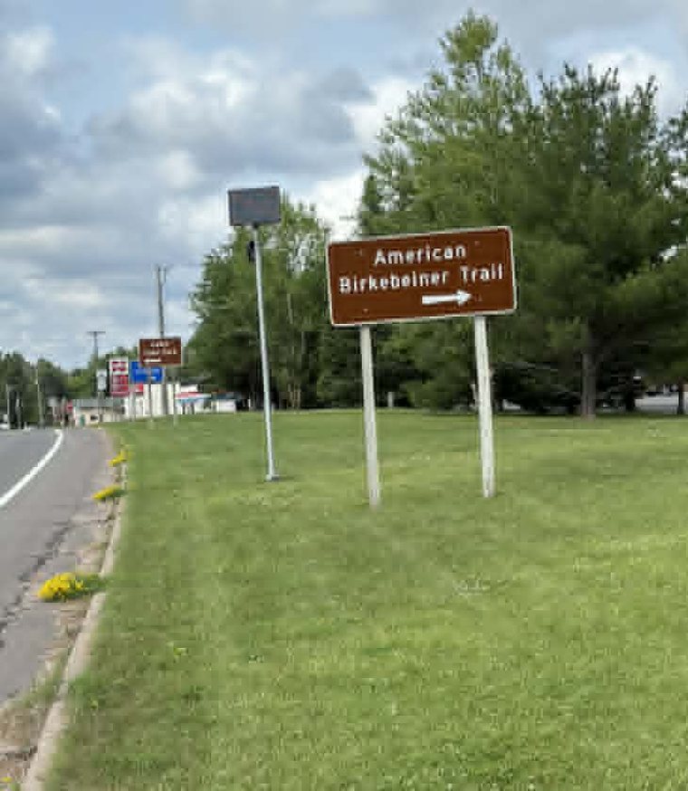 A solar-powered Flock license-plate camera on a roadside pole along US-63 in Cable, Wisconsin, next to the American Birkebeiner Trail sign